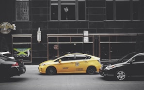A yellow taxi is parked on the street between two black cars in front of a large building with dark windows. The building has signage for the Hard Rock Hotel and another sign advertising Jamaican cuisine. A vertical light fixture decorates the facade, and there are no visible reflections in the windows.