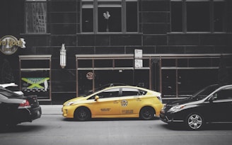 A yellow taxi is parked on the street between two black cars in front of a large building with dark windows. The building has signage for the Hard Rock Hotel and another sign advertising Jamaican cuisine. A vertical light fixture decorates the facade, and there are no visible reflections in the windows.