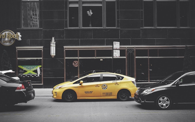 A yellow taxi is parked on the street between two black cars in front of a large building with dark windows. The building has signage for the Hard Rock Hotel and another sign advertising Jamaican cuisine. A vertical light fixture decorates the facade, and there are no visible reflections in the windows.