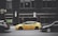 A yellow taxi is parked on the street between two black cars in front of a large building with dark windows. The building has signage for the Hard Rock Hotel and another sign advertising Jamaican cuisine. A vertical light fixture decorates the facade, and there are no visible reflections in the windows.