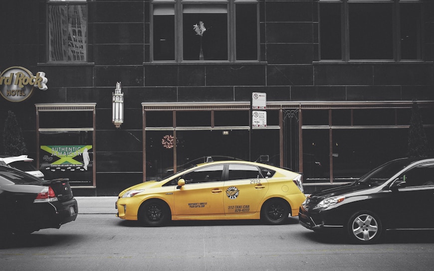 A yellow taxi is parked on the street between two black cars in front of a large building with dark windows. The building has signage for the Hard Rock Hotel and another sign advertising Jamaican cuisine. A vertical light fixture decorates the facade, and there are no visible reflections in the windows.