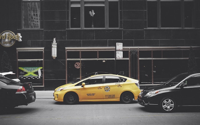 A yellow taxi is parked on the street between two black cars in front of a large building with dark windows. The building has signage for the Hard Rock Hotel and another sign advertising Jamaican cuisine. A vertical light fixture decorates the facade, and there are no visible reflections in the windows.