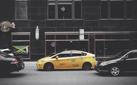 A yellow taxi is parked on the street between two black cars in front of a large building with dark windows. The building has signage for the Hard Rock Hotel and another sign advertising Jamaican cuisine. A vertical light fixture decorates the facade, and there are no visible reflections in the windows.