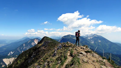 A lone hiker pauses atop a rocky summit, gazing over lush green valleys under a clear blue sky.