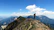 A smiling hiker standing on a mountain ridge overlooking lush green valleys under a clear blue sky.