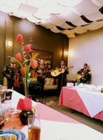 A group of musicians dressed in traditional outfits perform in a cozy indoor setting. Tulips in a vase adorn the table, while the ceiling is covered with fabric drapes creating a warm ambiance. Plates and drinks are set on the table, suggesting a festive gathering.