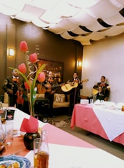 A group of musicians dressed in traditional outfits perform in a cozy indoor setting. Tulips in a vase adorn the table, while the ceiling is covered with fabric drapes creating a warm ambiance. Plates and drinks are set on the table, suggesting a festive gathering.
