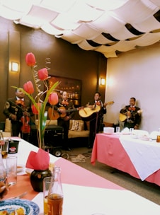 A group of musicians dressed in traditional outfits perform in a cozy indoor setting. Tulips in a vase adorn the table, while the ceiling is covered with fabric drapes creating a warm ambiance. Plates and drinks are set on the table, suggesting a festive gathering.
