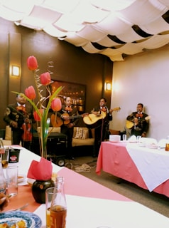 A group of musicians dressed in traditional outfits perform in a cozy indoor setting. Tulips in a vase adorn the table, while the ceiling is covered with fabric drapes creating a warm ambiance. Plates and drinks are set on the table, suggesting a festive gathering.