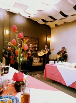 A group of musicians dressed in traditional outfits perform in a cozy indoor setting. Tulips in a vase adorn the table, while the ceiling is covered with fabric drapes creating a warm ambiance. Plates and drinks are set on the table, suggesting a festive gathering.