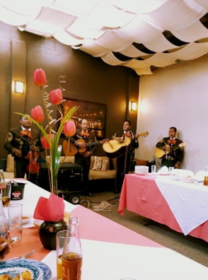 A group of musicians dressed in traditional outfits perform in a cozy indoor setting. Tulips in a vase adorn the table, while the ceiling is covered with fabric drapes creating a warm ambiance. Plates and drinks are set on the table, suggesting a festive gathering.