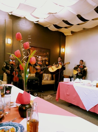 A group of musicians dressed in traditional outfits perform in a cozy indoor setting. Tulips in a vase adorn the table, while the ceiling is covered with fabric drapes creating a warm ambiance. Plates and drinks are set on the table, suggesting a festive gathering.