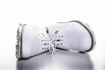 A pair of white leather golf shoes laid on their sides with soft spikes visible on the soles. The shoes have intricate stitching and laces, with branding visible on the side.