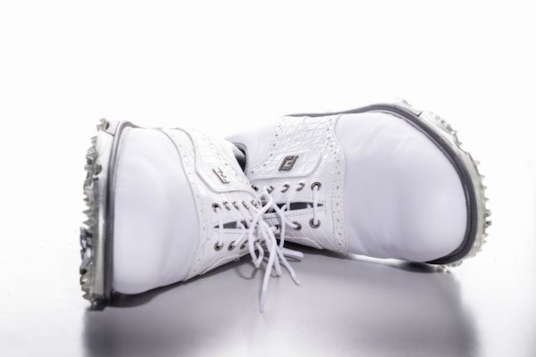 A pair of white leather golf shoes laid on their sides with soft spikes visible on the soles. The shoes have intricate stitching and laces, with branding visible on the side.