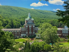 A large, grand hotel with a distinctive tower is nestled amidst lush green trees and rolling hills. The building features a red brick facade with white accents and numerous windows. Surrounding the hotel are well-manicured gardens and pathways, all under a clear blue sky with a few fluffy clouds.