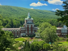 A large, grand hotel with a distinctive tower is nestled amidst lush green trees and rolling hills. The building features a red brick facade with white accents and numerous windows. Surrounding the hotel are well-manicured gardens and pathways, all under a clear blue sky with a few fluffy clouds.