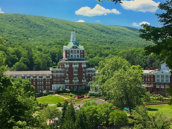 A large, grand hotel with a distinctive tower is nestled amidst lush green trees and rolling hills. The building features a red brick facade with white accents and numerous windows. Surrounding the hotel are well-manicured gardens and pathways, all under a clear blue sky with a few fluffy clouds.
