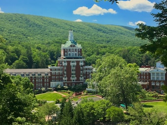 A large, grand hotel with a distinctive tower is nestled amidst lush green trees and rolling hills. The building features a red brick facade with white accents and numerous windows. Surrounding the hotel are well-manicured gardens and pathways, all under a clear blue sky with a few fluffy clouds.