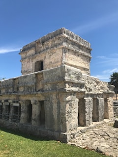 An ancient stone structure with rectangular stone blocks and columns set against a clear blue sky. The building features small openings and carvings, indicative of historical architecture.