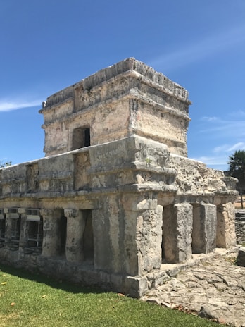 An ancient stone structure with rectangular stone blocks and columns set against a clear blue sky. The building features small openings and carvings, indicative of historical architecture.