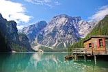 A picturesque wooden cabin sits on the edge of a tranquil turquoise lake, surrounded by towering, rugged mountains and lush green forests under a clear blue sky. A small boat is moored by the cabin's dock.