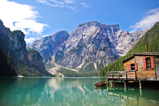 A picturesque wooden cabin sits on the edge of a tranquil turquoise lake, surrounded by towering, rugged mountains and lush green forests under a clear blue sky. A small boat is moored by the cabin's dock.