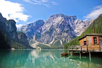 A picturesque wooden cabin sits on the edge of a tranquil turquoise lake, surrounded by towering, rugged mountains and lush green forests under a clear blue sky. A small boat is moored by the cabin's dock.