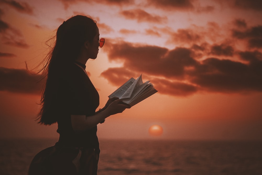 Woman with sunglasses holding book with a sunrise over ocean as the backdrop