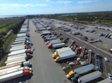 An aerial view captures a large parking lot filled with numerous semi-trucks and trailers arranged in rows. The surrounding area includes lush green fields and a body of water in the distance. The sky is clear with a few clouds, suggesting a bright day.