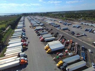 An aerial view captures a large parking lot filled with numerous semi-trucks and trailers arranged in rows. The surrounding area includes lush green fields and a body of water in the distance. The sky is clear with a few clouds, suggesting a bright day.