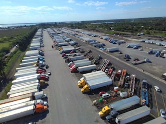 An aerial view captures a large parking lot filled with numerous semi-trucks and trailers arranged in rows. The surrounding area includes lush green fields and a body of water in the distance. The sky is clear with a few clouds, suggesting a bright day.