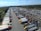 An aerial view captures a large parking lot filled with numerous semi-trucks and trailers arranged in rows. The surrounding area includes lush green fields and a body of water in the distance. The sky is clear with a few clouds, suggesting a bright day.