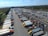 An aerial view captures a large parking lot filled with numerous semi-trucks and trailers arranged in rows. The surrounding area includes lush green fields and a body of water in the distance. The sky is clear with a few clouds, suggesting a bright day.