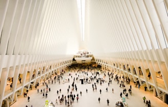 An expansive, futuristic indoor space with a vast open floor and high white ribbed ceilings converging at a vanishing point. Numerous people are walking or standing, with stalls and small structures scattered around the area, suggestive of a shopping or transportation hub.