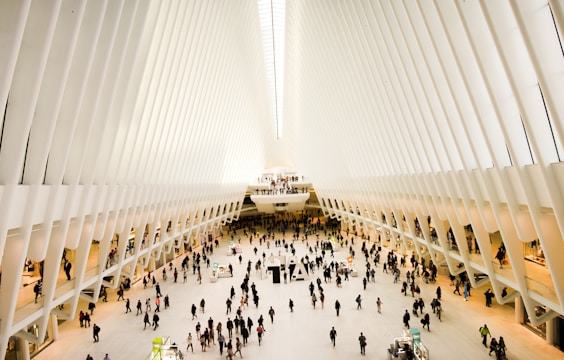 An expansive, futuristic indoor space with a vast open floor and high white ribbed ceilings converging at a vanishing point. Numerous people are walking or standing, with stalls and small structures scattered around the area, suggestive of a shopping or transportation hub.