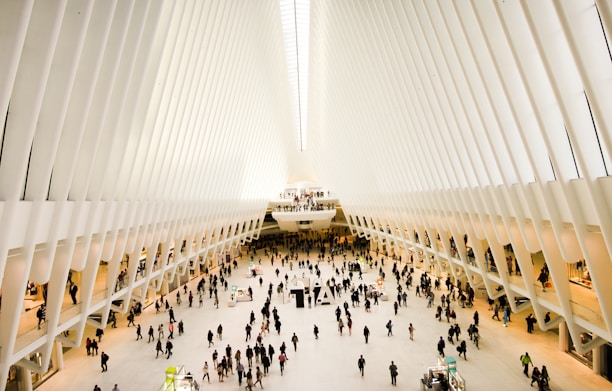 An expansive, futuristic indoor space with a vast open floor and high white ribbed ceilings converging at a vanishing point. Numerous people are walking or standing, with stalls and small structures scattered around the area, suggestive of a shopping or transportation hub.