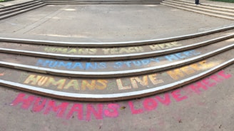 Colorful chalk messages on concrete steps convey sentiments like 'Humans teach here,' 'Humans study here,' 'Humans live here,' and 'Humans love here.' The steps are neatly aligned in a semi-circle pattern, with each message written in different colors.