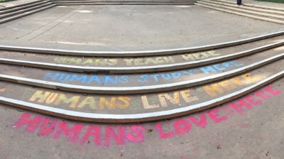Colorful chalk messages on concrete steps convey sentiments like 'Humans teach here,' 'Humans study here,' 'Humans live here,' and 'Humans love here.' The steps are neatly aligned in a semi-circle pattern, with each message written in different colors.