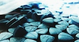A close-up view of numerous blue triangular tablets scattered and stacked together, some with visible imprints. The lighting creates a metallic sheen on the tablets, and there is a slight blur in the background.