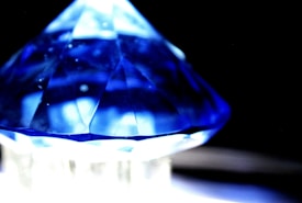 A faceted blue crystal or gemstone with a pyramid-like shape is positioned dramatically against a dark background. The light plays off the facets, creating a vibrant mix of blue and white reflections.
