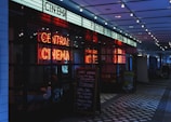 A dimly lit cinema entrance featuring a bright neon sign that reads 'Central Cinema'. The entrance includes a promotional chalkboard on the right listing various food and drink offerings. Overhead, string lights add to the atmospheric illumination.