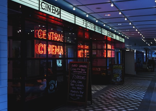 A dimly lit cinema entrance featuring a bright neon sign that reads 'Central Cinema'. The entrance includes a promotional chalkboard on the right listing various food and drink offerings. Overhead, string lights add to the atmospheric illumination.