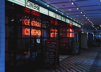 A dimly lit cinema entrance featuring a bright neon sign that reads 'Central Cinema'. The entrance includes a promotional chalkboard on the right listing various food and drink offerings. Overhead, string lights add to the atmospheric illumination.