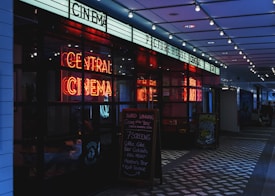 A dimly lit cinema entrance featuring a bright neon sign that reads 'Central Cinema'. The entrance includes a promotional chalkboard on the right listing various food and drink offerings. Overhead, string lights add to the atmospheric illumination.