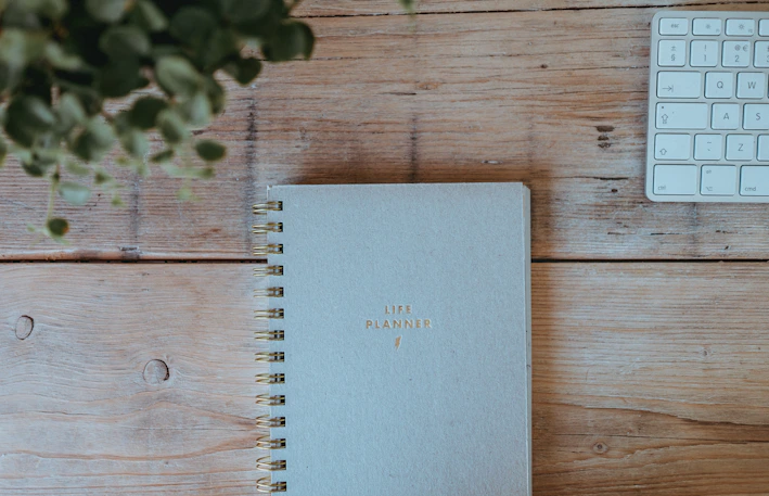 A light blue spiral-bound planner with the words 'Life Planner' embossed in gold sits on a wooden table. To the right is part of a white computer keyboard, and a leafy green plant is partially visible on the left.