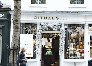 A storefront with the name 'Rituals' adorned above the entrance. The entrance is decorated with white floral arrangements. Inside, shelves with various products can be seen, accompanied by soft lighting. A woman with blonde hair, wearing a black jacket, is walking past on the left side of the image. A tree is also visible in the foreground to the left of the store.
