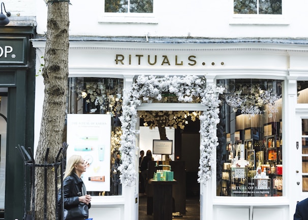 A storefront with the name 'Rituals' adorned above the entrance. The entrance is decorated with white floral arrangements. Inside, shelves with various products can be seen, accompanied by soft lighting. A woman with blonde hair, wearing a black jacket, is walking past on the left side of the image. A tree is also visible in the foreground to the left of the store.