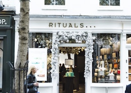 A storefront with the name 'Rituals' adorned above the entrance. The entrance is decorated with white floral arrangements. Inside, shelves with various products can be seen, accompanied by soft lighting. A woman with blonde hair, wearing a black jacket, is walking past on the left side of the image. A tree is also visible in the foreground to the left of the store.