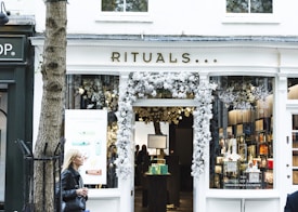 A storefront with the name 'Rituals' adorned above the entrance. The entrance is decorated with white floral arrangements. Inside, shelves with various products can be seen, accompanied by soft lighting. A woman with blonde hair, wearing a black jacket, is walking past on the left side of the image. A tree is also visible in the foreground to the left of the store.