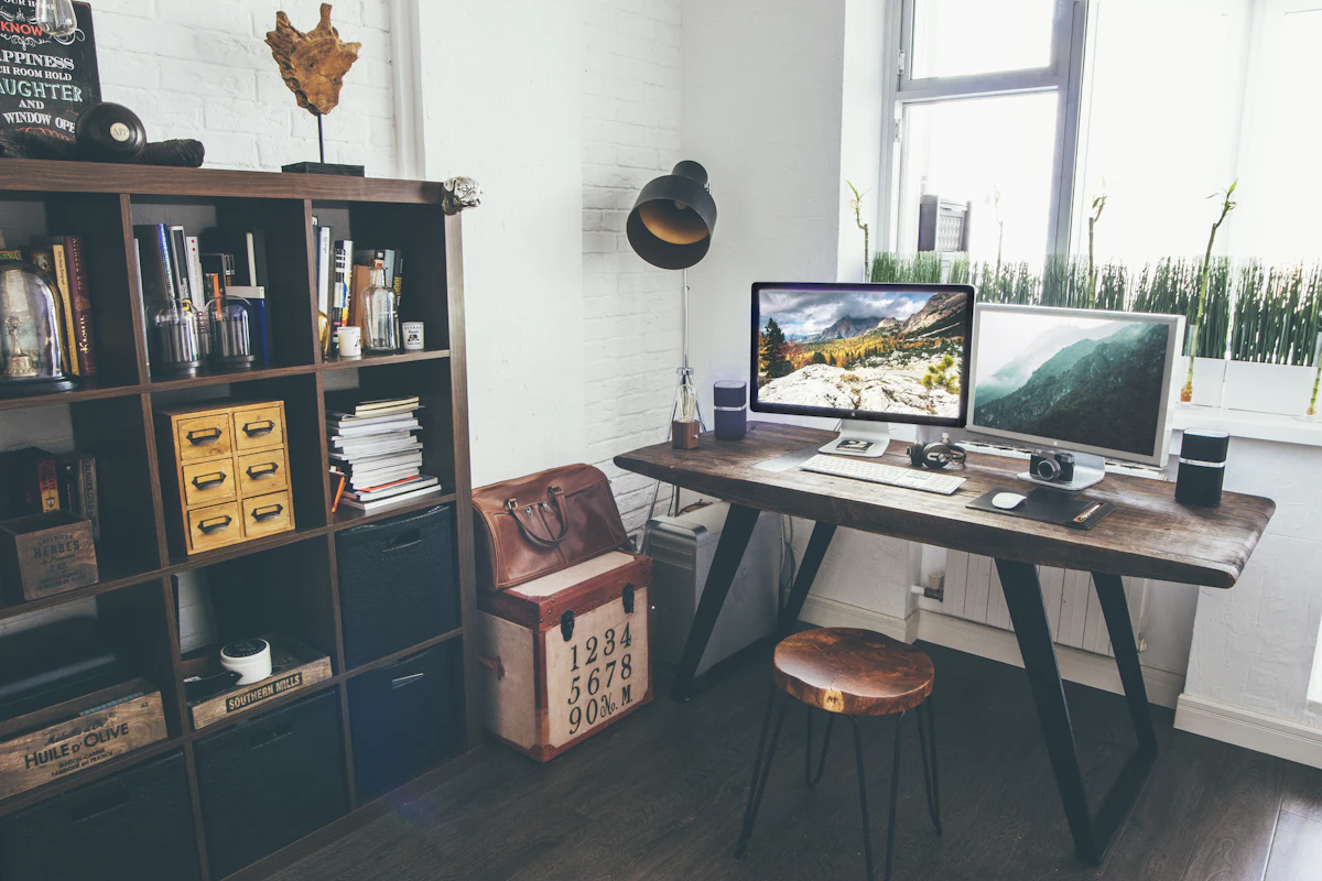 Un bureau à domicile avec un plateau en bois sur piètement noir, un iMac et un second écran affichant des photos de paysage, un sac en cuir sur une caisse numérotée, un tabouret rond en cuir et à gauche une étagère pleine de livres et de boîtes